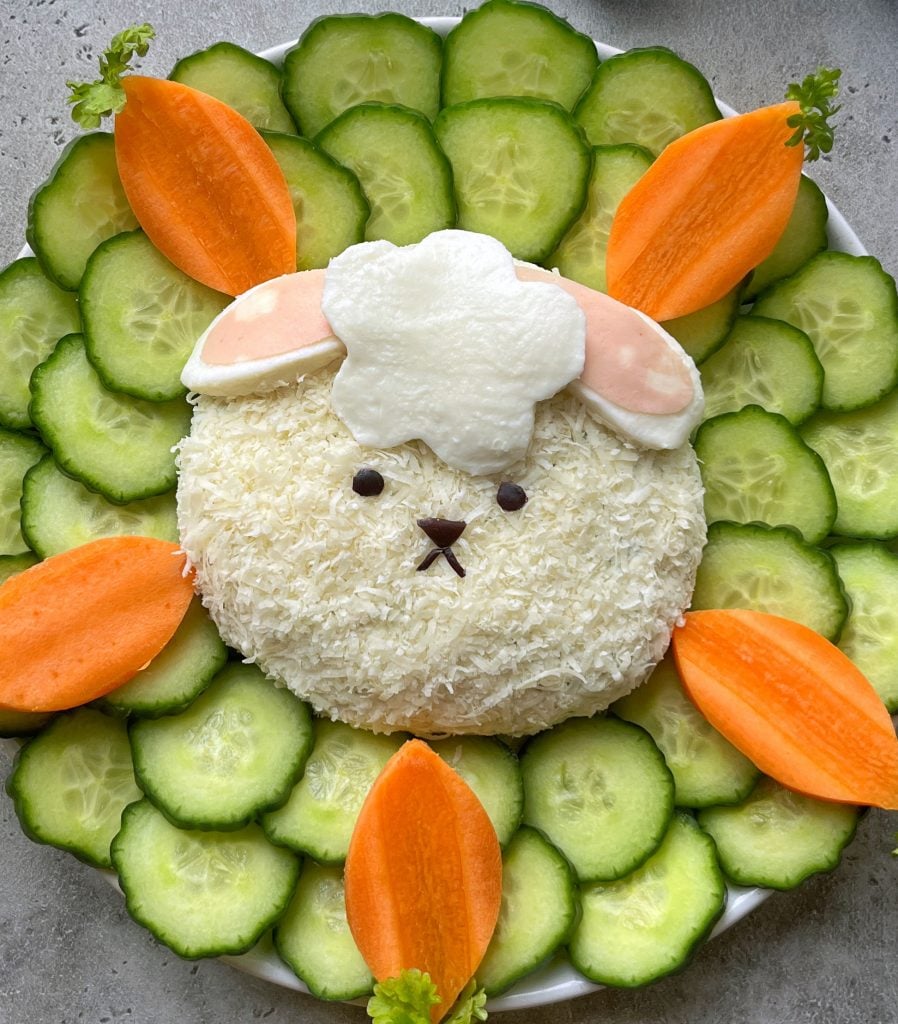 A cute Easter Cheese Ball shaped like a bunny, topped with white cheese and sliced ham, is surrounded by cucumber slices and carrot pieces arranged in a flower pattern on a white plate.