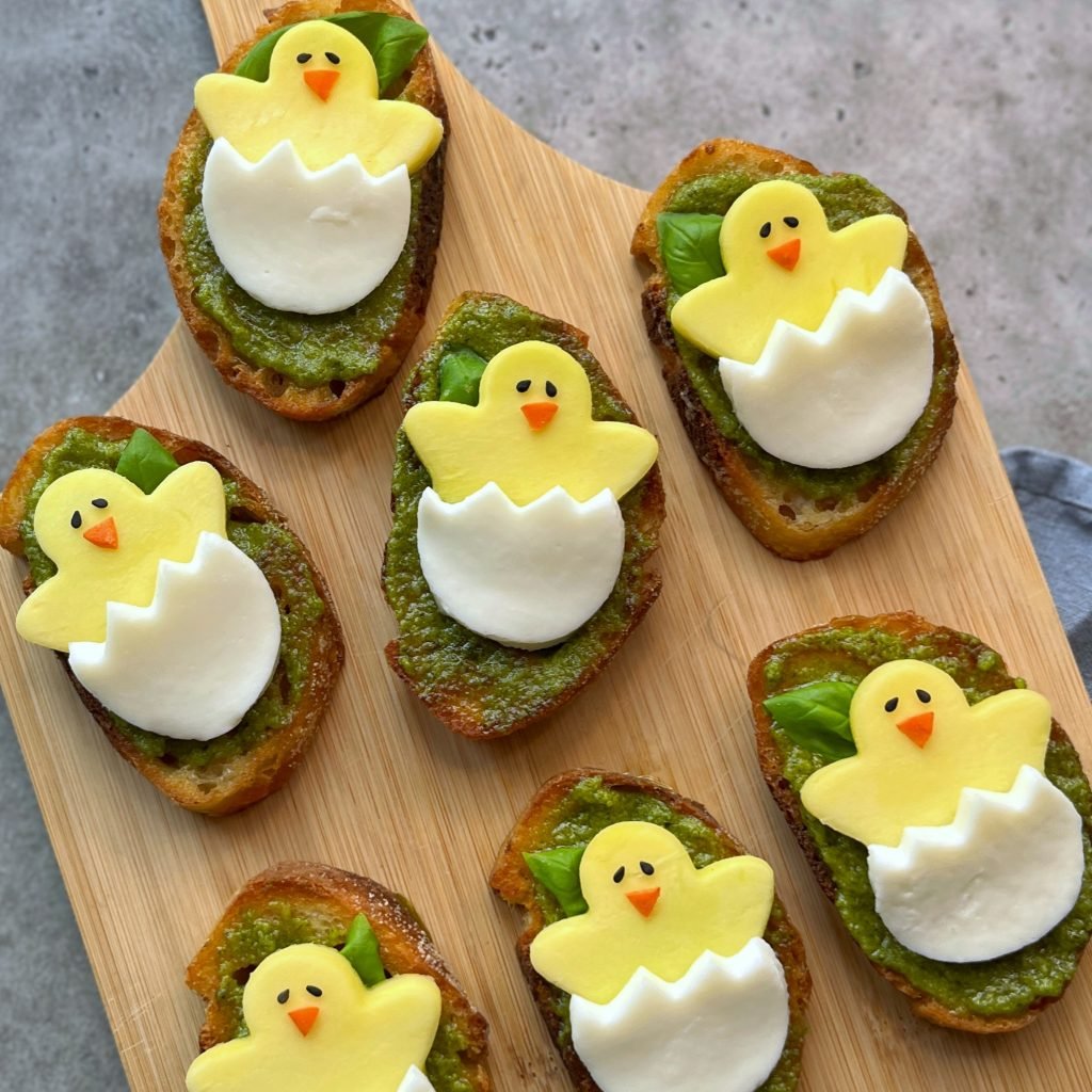 Sliced bread topped with green pesto, mozzarella, and mango cut into the shape of yellow chicks hatching from eggs arranged on a wooden board.