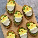 Sliced bread topped with green pesto, mozzarella, and cheese cut into the shape of yellow chicks hatching from eggs makes these adorable bites perfect for fun food recipes, arranged beautifully on a wooden board.