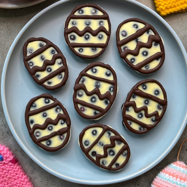 A plate with seven oval-shaped, white chocolate bites decorated like Easter eggs with dark chocolate zigzag and dot patterns, and visible pieces of dried fruit or berries inside. Crocheted decorations are partially visible beside the plate.