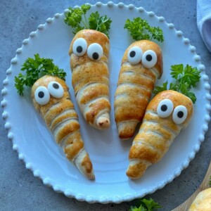 Four crescent roll snacks shaped like carrots, inspired by Easter Sausage Rolls, with googly eyes and parsley tops are arranged on a white plate, resembling cute, edible carrot creatures.
