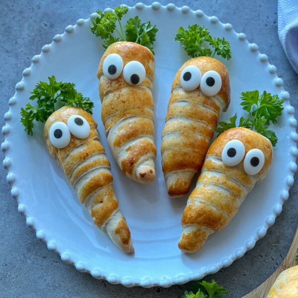 Four crescent roll snacks shaped like carrots, inspired by Easter Sausage Rolls, with googly eyes and parsley tops are arranged on a white plate, resembling cute, edible carrot creatures.