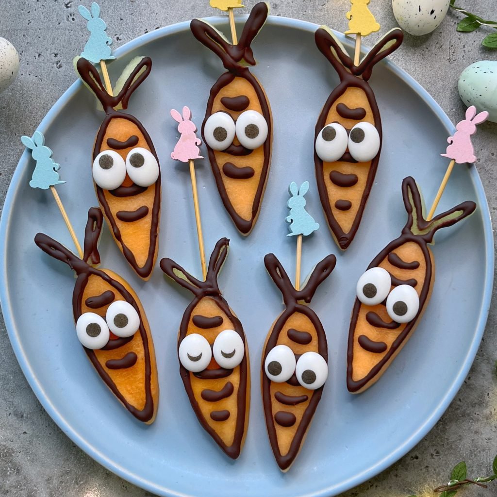 A closeup of seven carrot-shaped snacks made with melon decorated with chocolate outlines and candy eyes, arranged on a blue plate. 