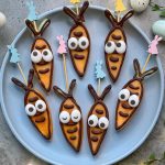 A plate of seven carrot-shaped cookies decorated with chocolate outlines and candy eyes, arranged in a circle. Some cookies are on sticks with pastel bunny toppers. Easter eggs are scattered around the plate, creating festive Easter bites.