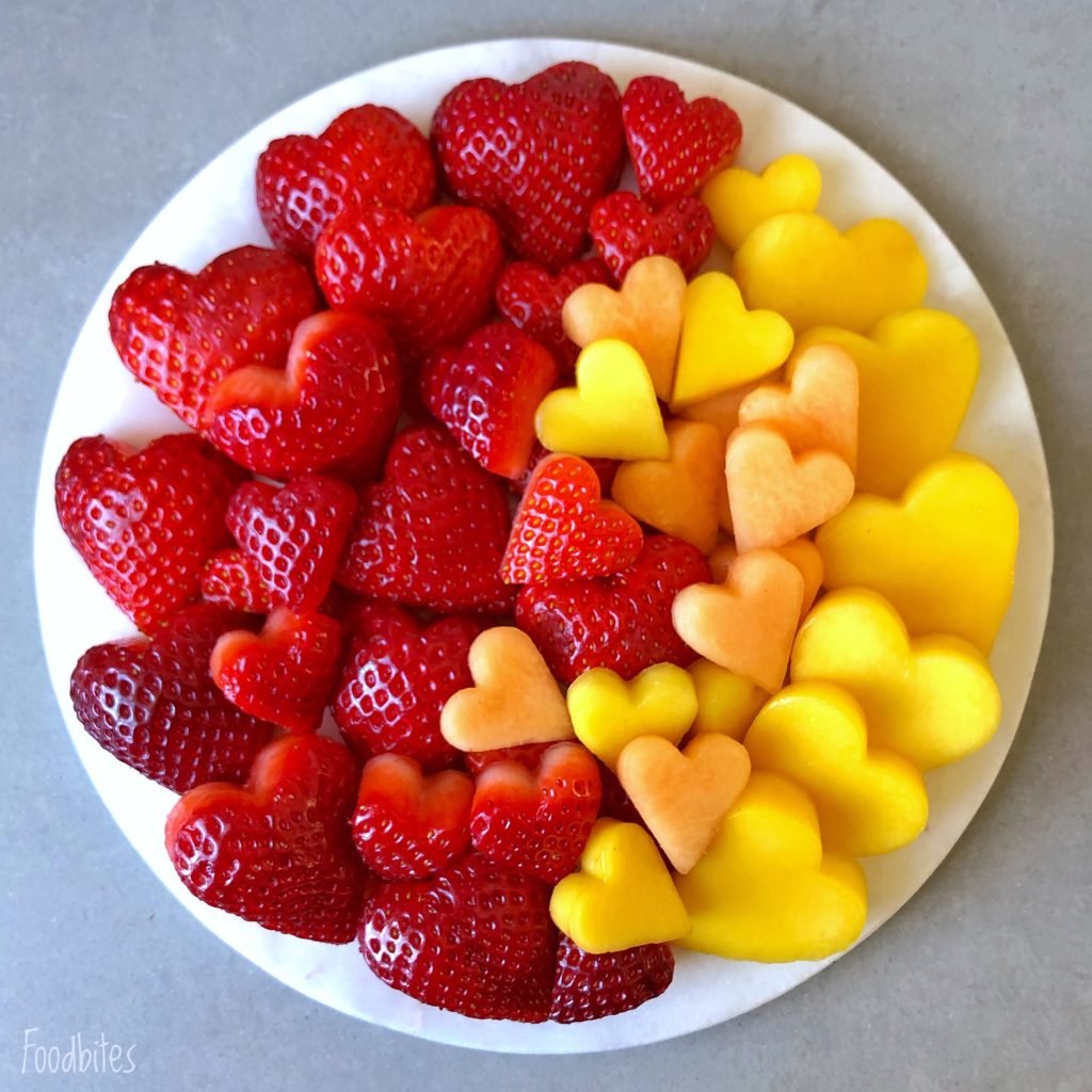 A round white plate with heart-shaped pieces of strawberries, cantaloupe, and mango arranged by color on a gray surface.