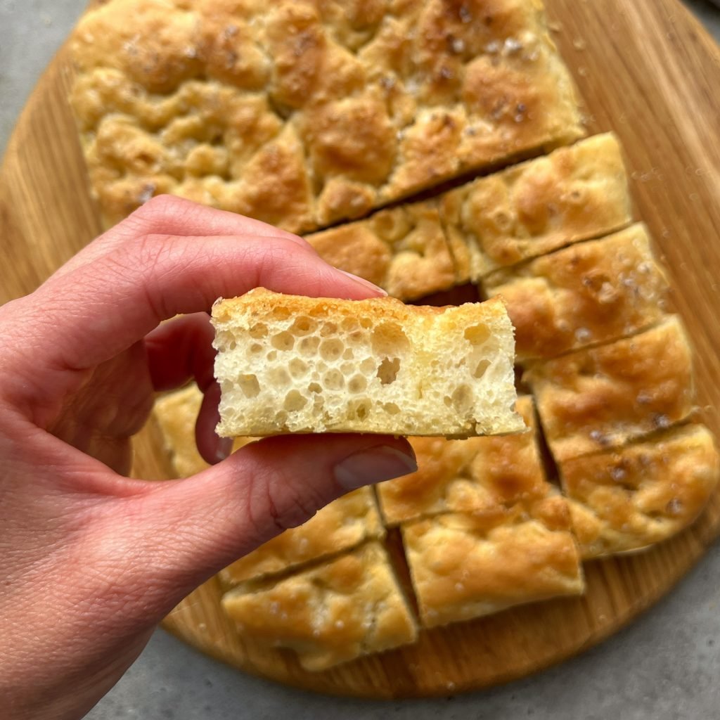 A hand holds a slice of airy, golden focaccia bread above a wooden board with more focaccia pieces. This delicious bite showcases the bread’s light, fluffy texture—perfect for adding to your favorite food recipes or sharing at casual gatherings.