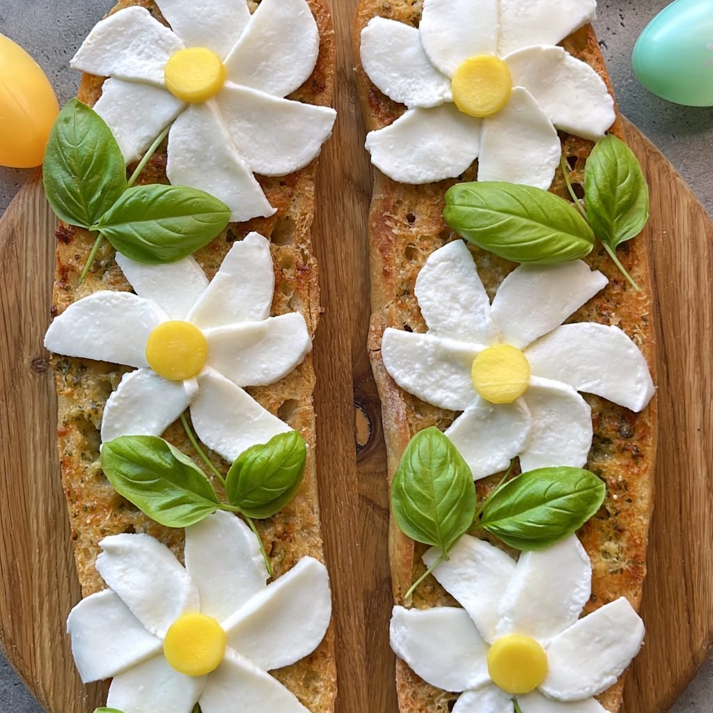 Two pieces of garlic bread on a wooden board are decorated with mozzarella slices as flower petals, mango centers, and fresh basil leaves as stems.
