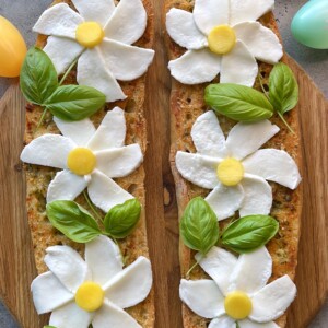 Two flatbreads on a wooden board are decorated with mozzarella slices as flower petals, yellow cheese centers, and fresh basil leaves as stems, creating a floral design—an inspiring bites idea for festive food recipes. Pastel-colored eggs sit at the edges.