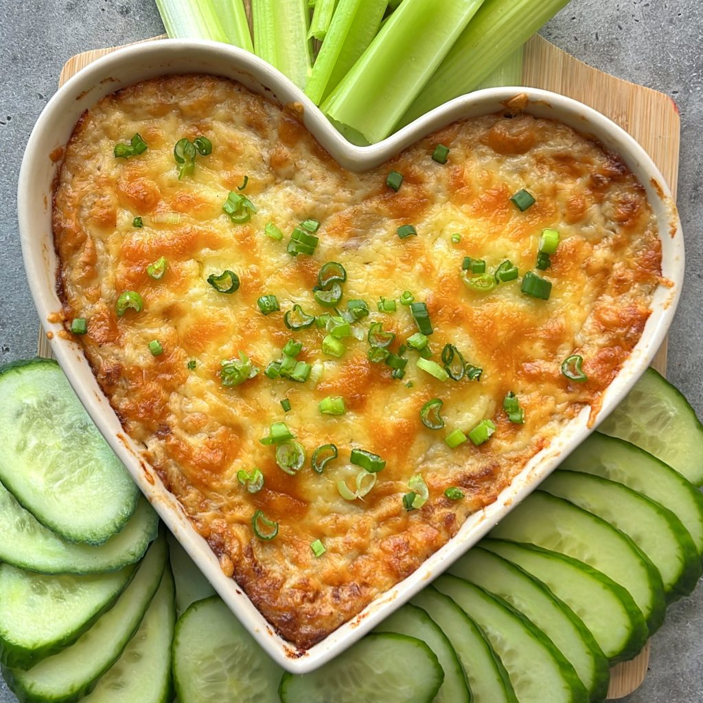 A heart-shaped dish filled with baked cheesy dip, garnished with chopped green onions. Slices of cucumber and sticks of celery are arranged around the dish on a wooden board for healthy bites.