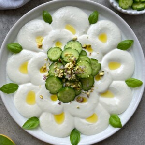 A creamy white feta cheese dip is arranged in a flower pattern, topped with cucumber slices in the center, drizzled with olive oil, chopped pistachios, red onion, and garnished with fresh basil leaves.