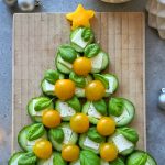A festive arrangement of cucumber slices, mozzarella, basil leaves, and yellow cherry tomatoes shaped like a Christmas tree on a wooden cutting board, topped with star-shaped yellow cheese—perfect for health-conscious holiday bites.