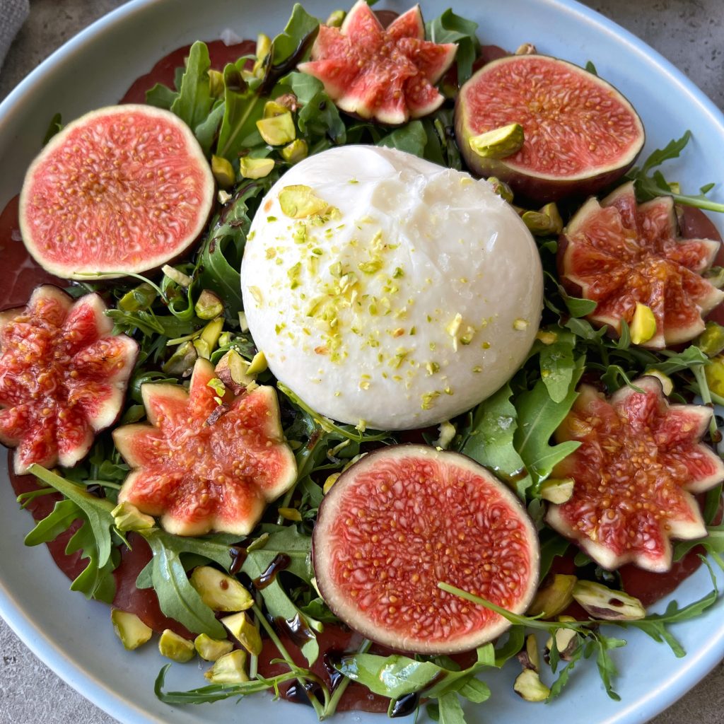 A plate of fresh arugula topped with whole burrata cheese, surrounded by decorative fig slices, sprinkled with pistachios, and drizzled with balsamic glaze—one of those irresistible bites perfect for health-minded food recipes.