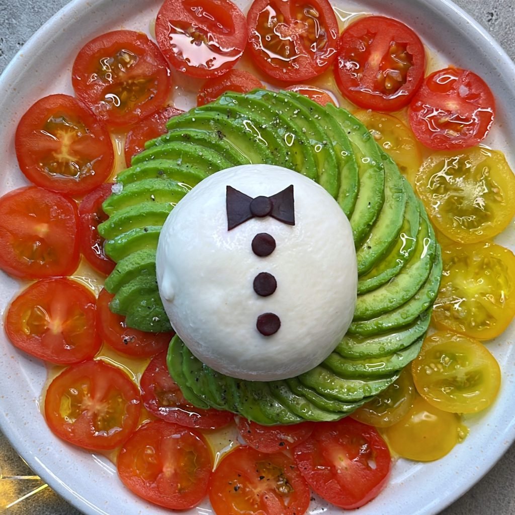 A plate with sliced red and yellow tomatoes arranged in a circle, topped with fanned-out avocado slices and a ball of mozzarella topped with a bowtie.