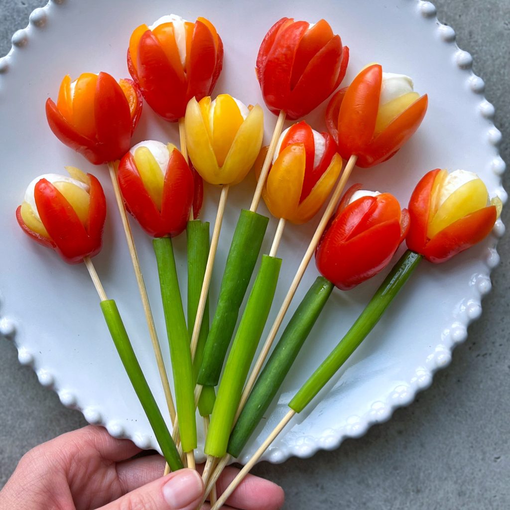 A hand holds a plate of skewered snacks arranged to look like tulip flowers, making a delightful mozzarella and tomato appetizer with red and yellow tomatoes, mozzarella centers, and green onions as stems, all served on a white plate.