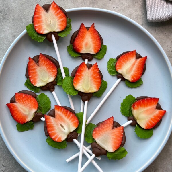 A plate displays lollipop sticks topped with halved strawberries and chocolate, arranged with green leaves to resemble edible flowers—perfect for healthy bites and creative food recipes.