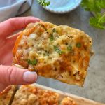 A hand holds a cheesy Focaccia Garlic Bread slice topped with melted cheese and chopped parsley. More pieces and fresh herbs are visible in the background on a light, textured surface.