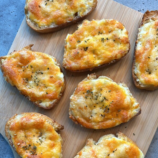 Slices of bread topped with melted cheese and herbs make for delicious bites, beautifully arranged on a wooden cutting board placed on a gray surface.