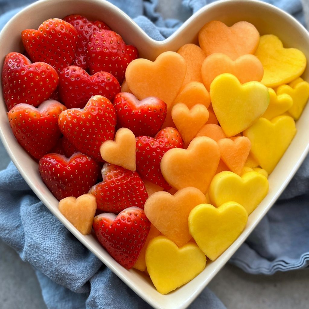 A heart-shaped bowl filled with heart-shaped pieces of strawberries, cantaloupe, and yellow fruit, arranged in a gradient from red to orange to yellow, sits on a light blue cloth.