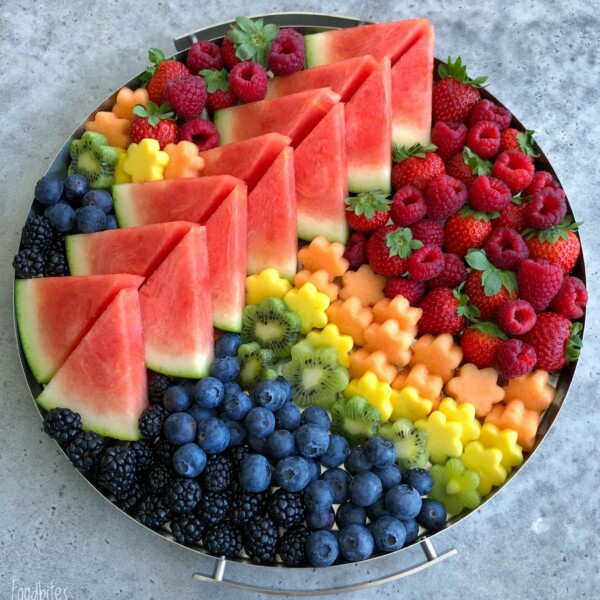 A round platter filled with a vibrant fruit medley—neatly arranged watermelon slices, raspberries, strawberries, blueberries, blackberries, kiwi slices, and flower-shaped pieces of cantaloupe and pineapple.