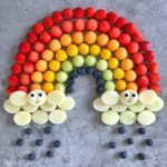 A rainbow made of rows of strawberries, cantaloupe, mango, kiwi, and blueberries, with clouds of sliced melon at both ends—each cloud features a smiling face. This colorful food dish turns fruit bites into edible art for health and happiness.