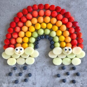 A rainbow made of rows of strawberries, cantaloupe, mango, kiwi, and blueberries, with clouds of sliced melon at both ends—each cloud features a smiling face. This colorful food dish turns fruit bites into edible art for health and happiness.