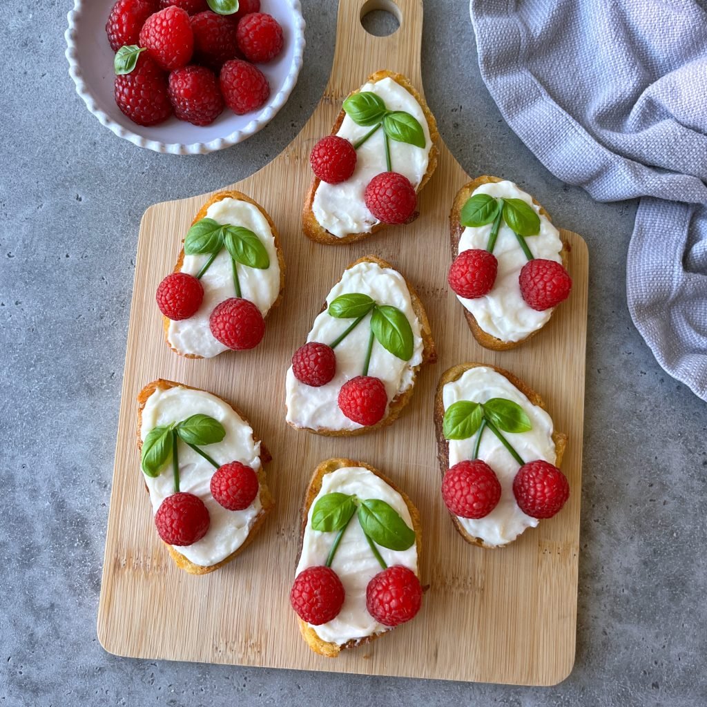 Seven pieces of goat cheese crostini, each topped with two raspberries and basil leaves to resemble cherries, are arranged on a wooden board. A bowl of raspberries and a gray cloth sit close by.