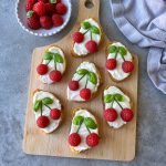 Seven pieces of goat cheese crostini, each topped with two raspberries and basil leaves to resemble cherries, are arranged on a wooden board. A bowl of raspberries and a gray cloth sit close by.