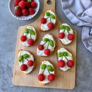 Seven pieces of goat cheese crostini, each topped with two raspberries and basil leaves to resemble cherries, are arranged on a wooden board. A bowl of raspberries and a gray cloth sit close by.