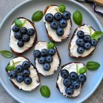 Six pieces of toast topped with goat cheese with honey and blueberries, arranged to resemble grape clusters, are garnished with fresh basil leaves on a light blue plate.