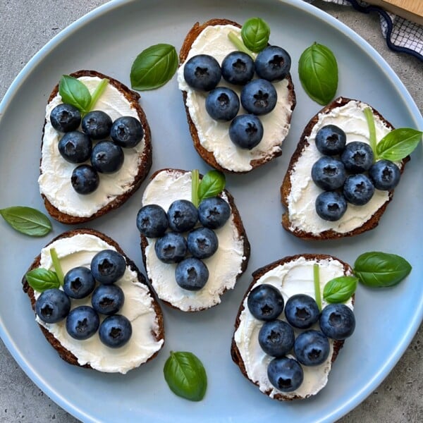 Six pieces of toast topped with goat cheese with honey and blueberries, arranged to resemble grape clusters, are garnished with fresh basil leaves on a light blue plate.