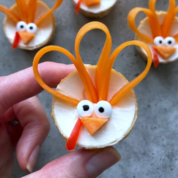 A hand holds a round cracker topped with a white spread, candy eyes, carrot loops for feathers, a triangle carrot beak, and red pepper wattle—crafted into a turkey face. These fun bites make festive additions to health-conscious food recipes.