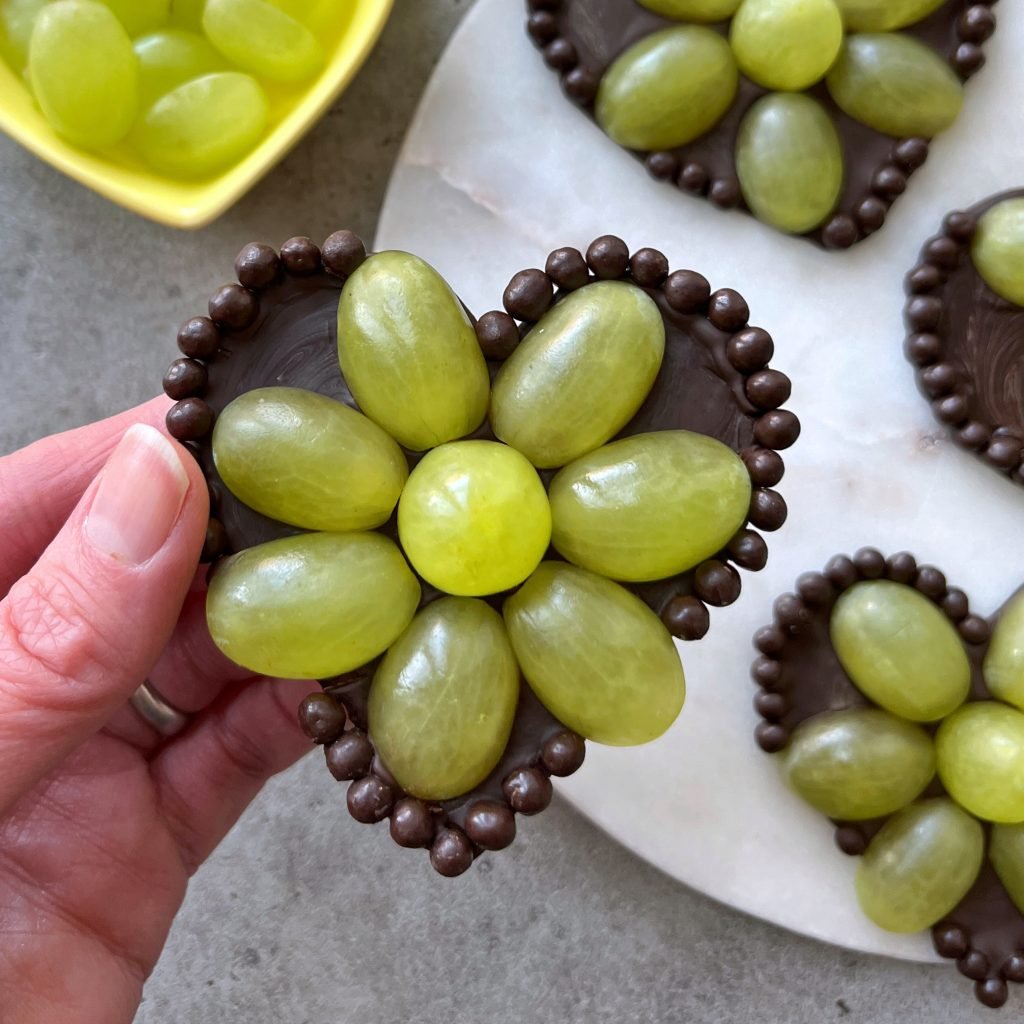 A hand holds a heart-shaped chocolate base decorated with green grapes arranged like flower petals and outlined with small round chocolates. More similar treats and a bowl of grapes appear in the background.