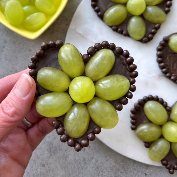 A hand holds a Grape Chocolate Heart—a heart-shaped chocolate base decorated with green grapes arranged like flower petals and outlined with small round chocolates. More similar treats and a bowl of grapes appear in the background.