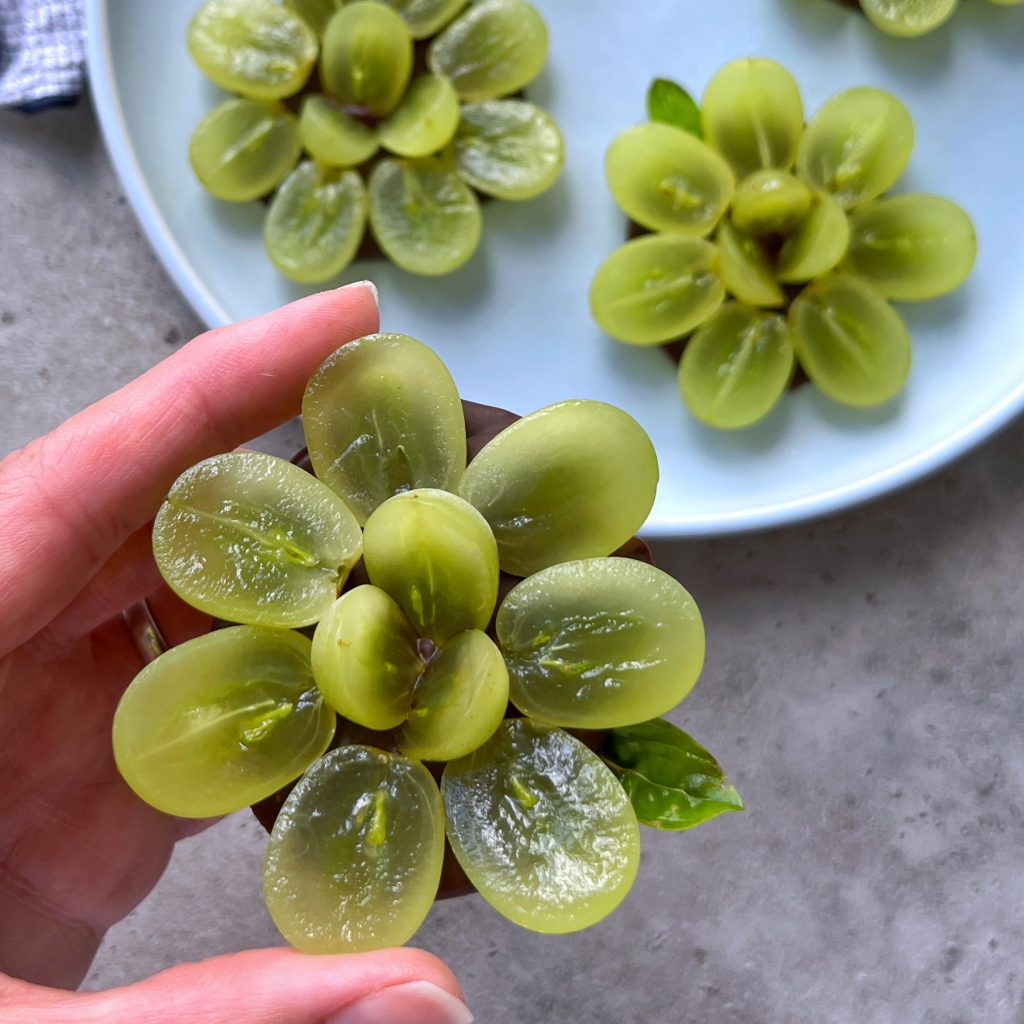 A hand holds a flower-shaped arrangement made from sliced green grapes, creating healthy bites displayed over a gray surface, with more grape flowers on a white plate in the background.