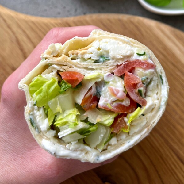 A hand holds a Greek Salad Wrap filled with lettuce, tomato, cucumber, and creamy white spread, with visible layers of soft tortilla on a wooden surface.