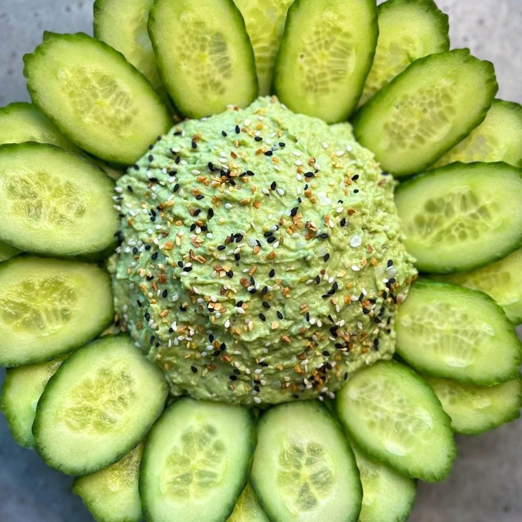 Closeup of a mound of creamy green dip topped with everything bagel seasoning is surrounded by overlapping cucumber slices, creating a fresh and vibrant dish.
