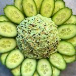 A mound of green dip topped with mixed seeds is surrounded by neatly arranged cucumber slices in a circular pattern on a gray surface, making for one of those health-focused dishes perfect for light bites.