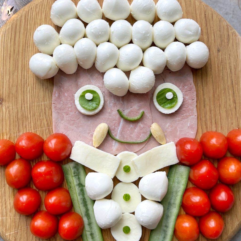 A playful Vampire portrait on a wooden board, using mozzarella balls for hair, ham for the face, blanched almonds for the fangs, cucumber and mozzarella for the body, and cherry tomatoes for the jacket.