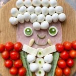 A playful food art portrait of a Vampire on a wooden board, using mozzarella balls for hair, ham for the face, cucumber and mozzarella for the body. His red jacket is made of cherry tomatoes.