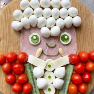 A playful food art portrait of a Vampire on a wooden board, using mozzarella balls for hair, ham for the face, cucumber and mozzarella for the body. His red jacket is made of cherry tomatoes.