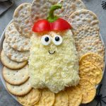 A Halloween-themed cheese ball shaped like a ghost, with shredded cheese, olive eyes, red bell pepper for a hat, and a smiling face—this delicious bite makes festive food recipes fun when served with assorted crackers on a plate.