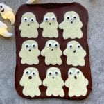 A slab of chocolate topped with nine white chocolate ghost shapes, each with candy eyes and different mouth expressions, arranged in a grid on a gray surface—perfect for fun food recipes or festive bites.