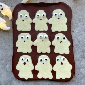 A slab of chocolate topped with nine white chocolate ghost shapes, each with candy eyes and different mouth expressions, arranged in a grid on a gray surface—perfect for fun food recipes or festive bites.
