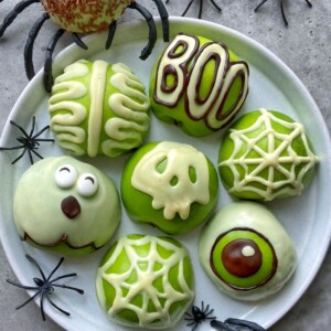 A plate of seven green Halloween-themed donuts—decorated as a brain, the word BOO, a monster face, a skull, two spiderwebs, and an eyeball—serves up spooky bites for your festive food recipes, with plastic spiders scattered around the plate.
