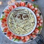 A round dessert bite decorated to look like a mummy with white chocolate drizzle as bandages and two candy eyes, surrounded by halved strawberries and plastic spiders on a white platter—perfect for spooky food recipes.