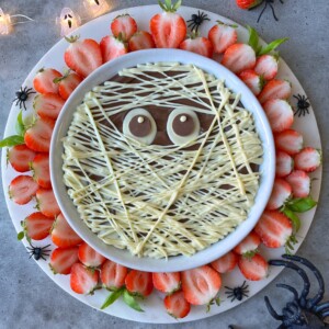 A round dessert bite decorated to look like a mummy with white chocolate drizzle as bandages and two candy eyes, surrounded by halved strawberries and plastic spiders on a white platter—perfect for spooky food recipes.