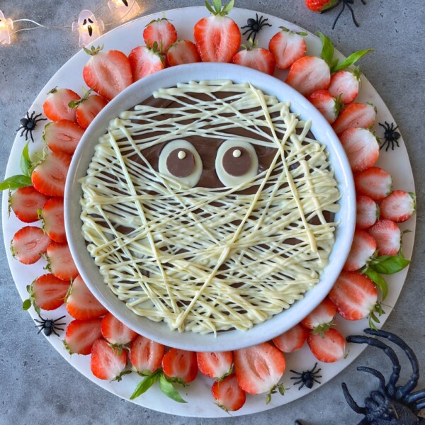 A round dessert bite decorated to look like a mummy with white chocolate drizzle as bandages and two candy eyes, surrounded by halved strawberries and plastic spiders on a white platter—perfect for spooky food recipes.