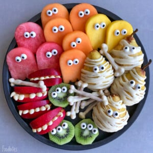 A plate of Halloween-themed bites features apple slices with teeth, kiwi slices, fruit cutouts with candy eyes, and pears wrapped like mummies with white icing and candy eyes—spooky snacks perfect for fun health-focused food recipes.