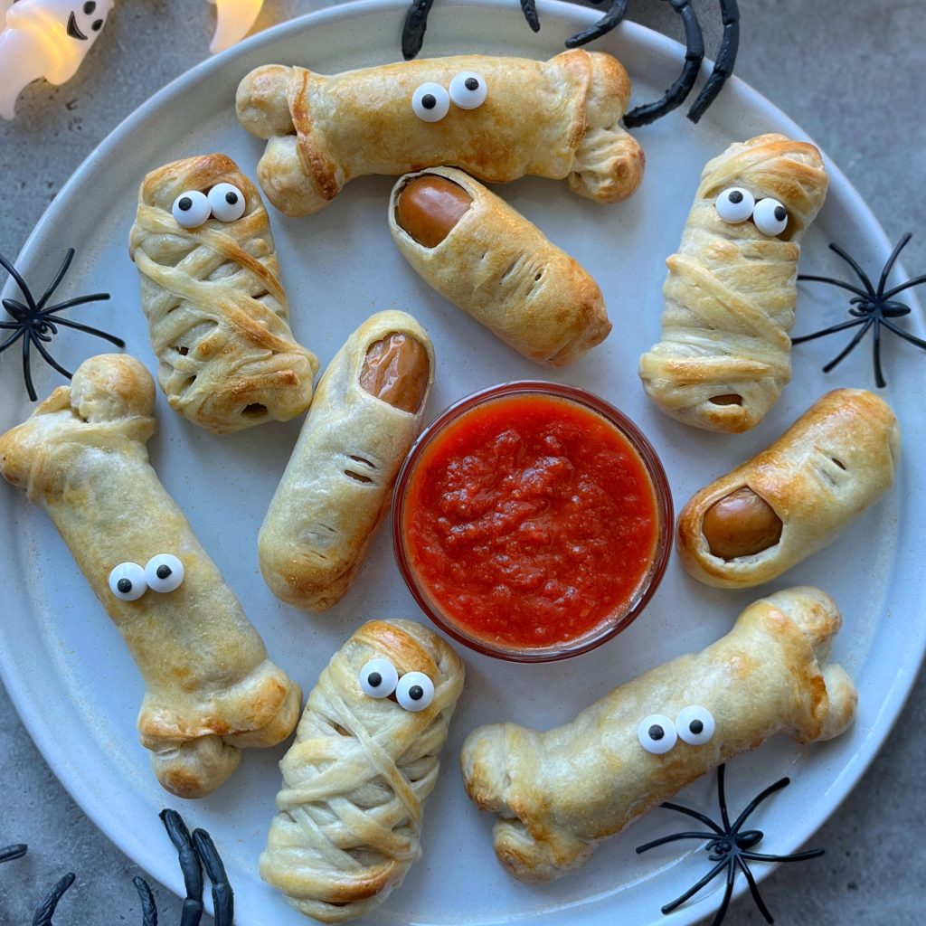 A plate of Halloween Sausage Rolls shaped like mummies and bones, made from pastry-wrapped sausages with candy eyes, surrounds a bowl of red dipping sauce. Plastic spiders are placed around the plate for a spooky touch.