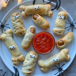 A plate of Halloween Sausage Rolls shaped like mummies and bones, made from pastry-wrapped sausages with candy eyes, surrounds a bowl of red dipping sauce. Plastic spiders are placed around the plate for a spooky touch.